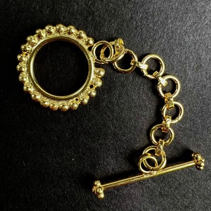 May include: A gold-colored toggle clasp with a circular design and a series of connecting rings. The clasp features a decorative beaded edge around the circular portion and a T-bar closure. The jewelry component is set against a black background.