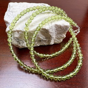 May include: A delicate necklace featuring small, round, light green beads. The beads are strung together, forming a multi-strand design. The necklace is displayed on a white, textured surface against a dark brown background.