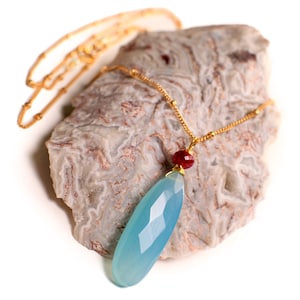 Può includere: Una collana con catena dorata con un ciondolo in pietra preziosa blu chiaro e un piccolo accento in pietra preziosa rossa. La collana è esposta su un pezzo di roccia.