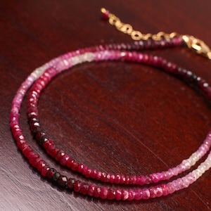 May include: A delicate necklace featuring small, faceted gemstones in shades of red, pink, and purple. The necklace has a gold-colored clasp and chain, and is displayed on a dark wooden surface.