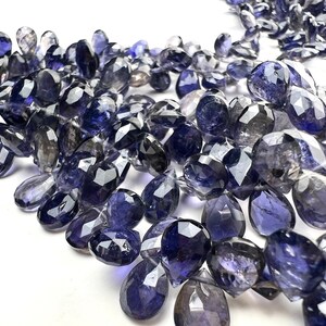 May include: A collection of faceted, teardrop-shaped iolite gemstones in varying shades of blue and gray. The gemstones are scattered across a white surface, showcasing their polished surfaces and internal clarity. The overall effect is one of elegance and natural beauty.