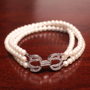 Puede incluir: Un collar de perlas de tres hileras con un cierre plateado decorado con brillantes transparentes.