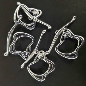 Op de afbeelding: Vijf zilveren metalen decoratieve haken met ingewikkelde ontwerpen. De haken zijn in een cirkelvormige opstelling op een zwarte achtergrond geplaatst.