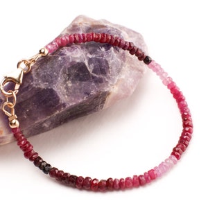 May include: A delicate bracelet made with a gradient of pink, red, and dark red gemstone beads. The bracelet has a gold clasp.