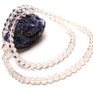 May include: A clear quartz crystal beaded necklace with a silver clasp. The necklace is laid out on a blue and purple stone.