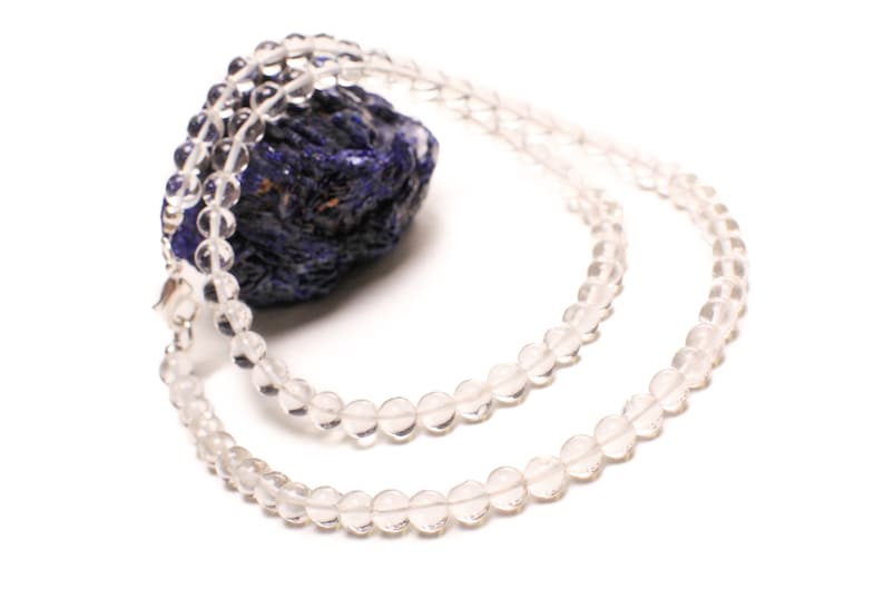 May include: A clear quartz crystal beaded necklace with a silver clasp. The necklace is laid out on a blue and purple stone.