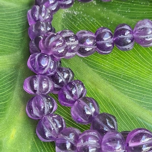 Può includere: Una collana di perle di ametista viola con una texture sfaccettata e rigata. Le perle sono infilate su un filo elastico trasparente e sono esposte su una foglia verde.