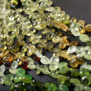 May include: A strand of green, yellow, and brown gemstone beads. The beads are faceted and have a pear shape. The beads are strung on a clear thread.