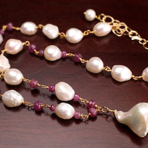 May include: A gold chain necklace featuring large, irregular white pearls and small, round, deep pink gemstones. The necklace has a pendant with a large, uniquely shaped pearl. The jewelry is displayed on a dark brown surface.