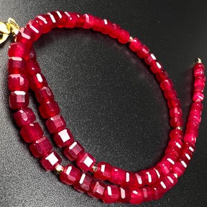 Puede incluir: Collar de piedras preciosas rojo vibrante con cierre dorado. El collar está compuesto por cuentas de rubí facetadas y cuadradas, ensartadas para crear un diseño de doble hebra. Las cuentas tienen una superficie pulida y reflectante. Longitud aproximada: 40 cm.
