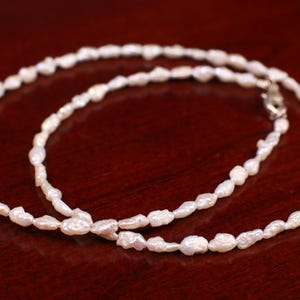 Könnte beinhalten: Eine weiße Perlenkette mit unregelmäßig geformten Perlen. Die Kette besteht aus kleinen, weißen Perlen, die auf einer silbernen Kette aufgereiht sind.