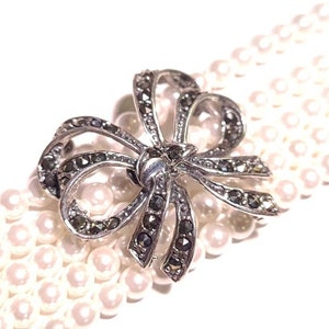 Op de afbeelding: Een zilveren en zwarte strik broche met een parelketting. De strik is gemaakt van zilver met zwarte stenen en de ketting is gemaakt van witte parels.