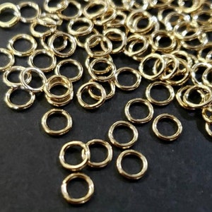 May include: A large pile of gold jump rings, a type of jewelry finding, on a black surface. The jump rings are small and round, and they are perfect for connecting different pieces of jewelry together.