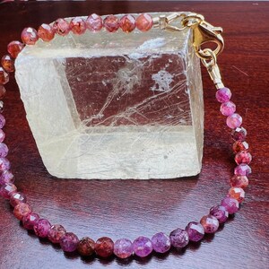 May include: A pink and red gemstone bracelet with a gold clasp. The bracelet is made of faceted beads and is strung on a gold chain.