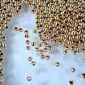 Puede incluir: Primer plano de un montón de cuentas redondas doradas. Las cuentas están esparcidas sobre una superficie blanca.