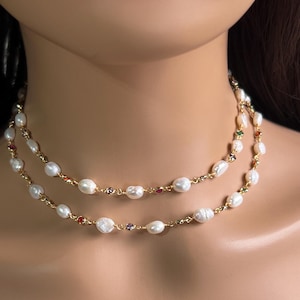 Puede incluir: Un collar de doble hebra con perlas de agua dulce blancas y pequeñas piedras preciosas de colores. El collar tiene una delicada cadena dorada. Las perlas tienen forma irregular, añadiendo un toque único a la joya.