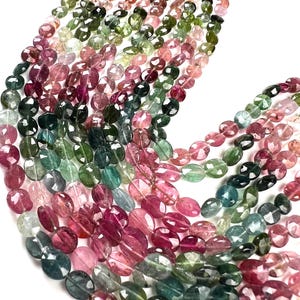 Puede incluir: Una colección de cuentas de turmalina ovaladas y facetadas en una variedad de colores que incluyen tonos rosa, verde, azul verdoso y amarillo. Las piedras preciosas translúcidas están dispuestas en múltiples hilos, mostrando un acabado pulido.