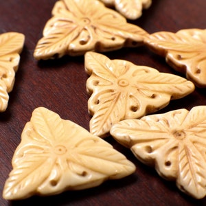 Può includere: Una collezione di sette ciondoli in osso intagliato a forma triangolare, di colore crema, con intricati motivi floreali.