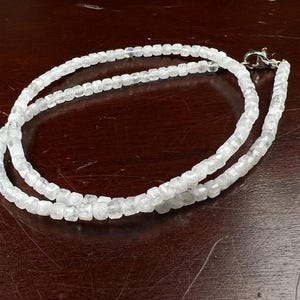 May include: A delicate, double-strand necklace composed of small, faceted, clear beads. The necklace has a silver-colored clasp and is displayed on a dark brown wooden surface. The beads appear to be evenly sized and reflect light.