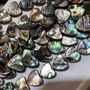 May include: A collection of iridescent abalone shell heart-shaped pendants. The shells are a mix of green, blue, and brown colors with a pearlescent sheen.