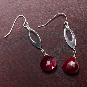 May include: A pair of silver dangle earrings with red gemstone drops. The earrings feature a simple oval shape with a cut-out design.