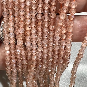 Könnte beinhalten: Nahaufnahme von Strängen facettierter, pfirsichfarbener Perlen. Die kleinen, runden Perlen haben ein funkelndes, kristallartiges Aussehen. Sie sind auf einem dünnen, orangefarbenen Faden aufgefädelt, was einen zarten Look erzeugt. Ideal für die Schmuckherstellung.