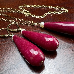 May include: A set of burgundy gemstone earrings and a necklace with a gold chain. Each piece features a teardrop-shaped gemstone with a pearl accent.