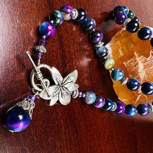 May include: A dark blue and purple gemstone beaded necklace with a silver butterfly charm and a toggle clasp. The necklace is on a wooden surface.