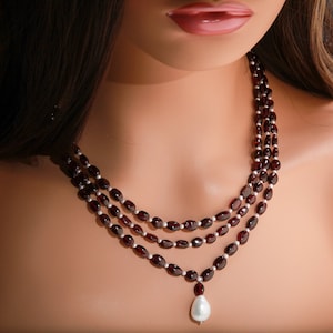 Può includere: Una collana a tre fili con un ciondolo di perla bianca grande. La collana è realizzata con perline di colore rosso scuro, di forma ovale.