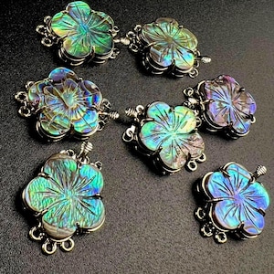 May include: Six iridescent abalone shell flower pendants with silver-colored metal accents. Each pendant features a carved floral design with a range of blues, greens, and purples. The pendants are arranged on a dark surface.