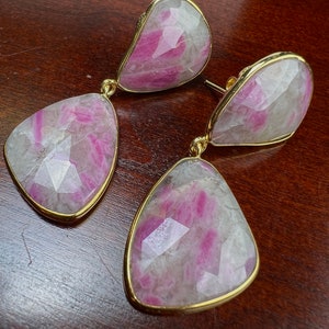 May include: A pair of gold earrings with pink and white gemstone drops. The earrings have a teardrop shape and are set in a gold frame.