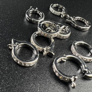 Könnte beinhalten: Nahaufnahme von mehreren silberfarbenen Schmuckverschlüssen mit funkelndem, edelsteinartigem Aussehen. Die Verschlüsse sind auf einer dunklen Oberfläche angeordnet, was ihr aufwendiges Design und ihre reflektierenden Eigenschaften zeigt. Die Verschlüsse sind wahrscheinlich für Halsketten oder Armbänder.