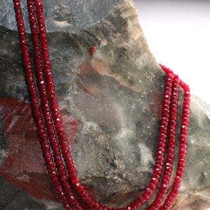 May include: A triple strand necklace made of red ruby beads. The necklace is draped over a rough, gray rock.