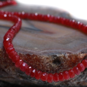 Puede incluir: Una hebra de cuentas de piedras preciosas rojas, probablemente rubíes o granates, enhebradas en un cordón fino. Las cuentas son facetadas y tienen un acabado brillante y pulido. Las cuentas están sobre una superficie rugosa y texturizada.