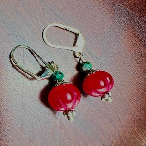 以下が含まれることがあります： 赤と緑のビーズが付いたイヤリングのペア。イヤリングはシルバーのフックが付いており、赤と緑のビーズを組み合わせて作られています。