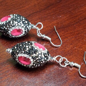 May include: A pair of dangle earrings with a silver hook. Each earring features a black and silver beaded oval shape with three pink oval accents. The earrings are on a wooden surface.