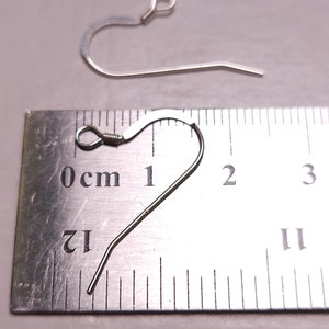 May include: Two silver-colored earring hooks are displayed against a light background. One hook is in focus, showing a curved design with a looped top and a straight, pointed bottom. The other hook is slightly out of focus. A ruler is in the background.