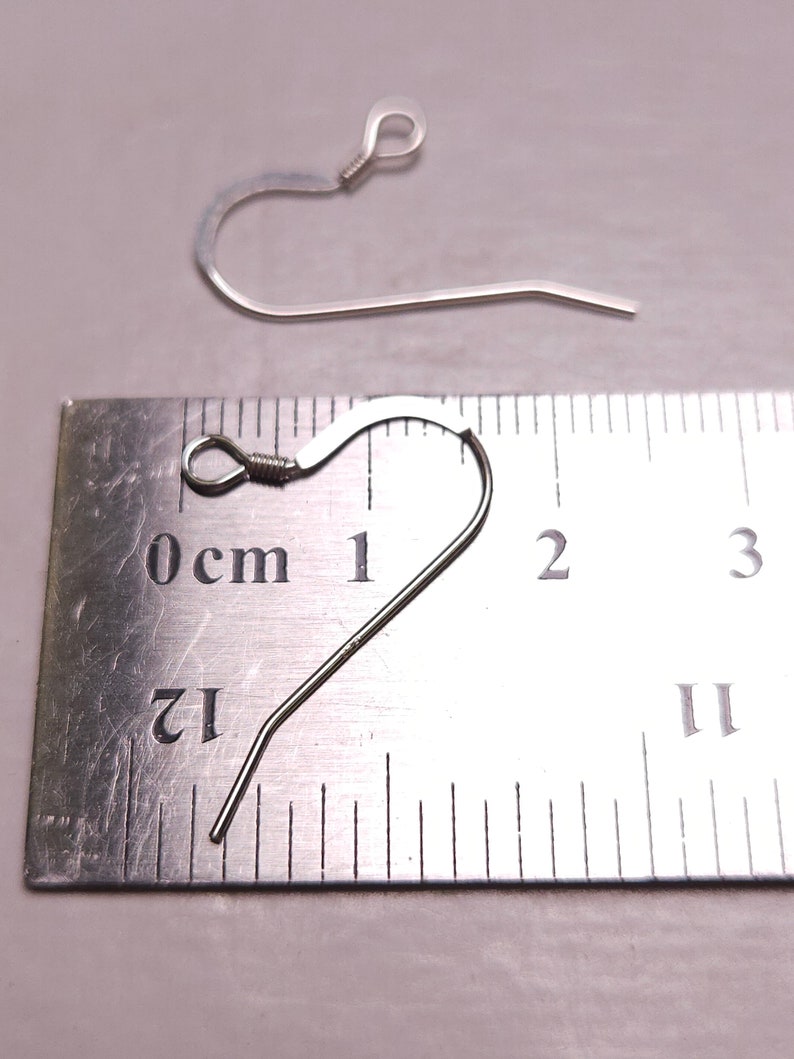 May include: Two silver-colored earring hooks are displayed against a light background. One hook is in focus, showing a curved design with a looped top and a straight, pointed bottom. The other hook is slightly out of focus. A ruler is in the background.