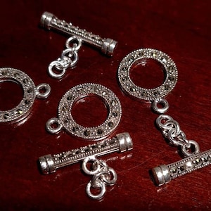 Könnte beinhalten: Fünf silberne Toggle-Verschlüsse mit einem runden, funkelnden Design. Jeder Verschluss hat eine kleine Kette, die an dem Ring befestigt ist.