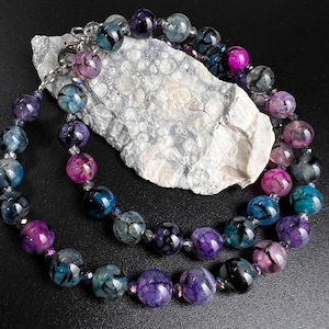 Puede incluir: Un collar de doble cadena hecho de cuentas de ágata. Las cuentas son una mezcla de morado, azul y negro con un patrón de remolino. El collar está sobre una roca blanca con un fondo negro.