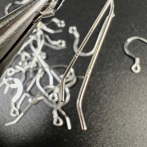 May include: Close-up of silver-colored earring components and jewelry-making tools. The image shows several earring hooks, ear wires, and a pair of ear climbers. A pair of pliers is visible, suggesting the process of jewelry assembly.