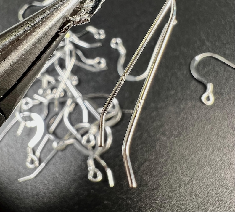 May include: Close-up of silver-colored earring components and jewelry-making tools. The image shows several earring hooks, ear wires, and a pair of ear climbers. A pair of pliers is visible, suggesting the process of jewelry assembly.
