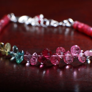 May include: A silver bracelet with a gradient of gemstones. The bracelet features green, blue, pink, and red gemstones.