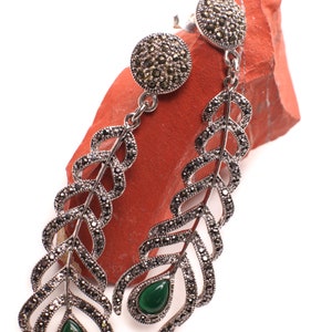 Puede incluir: Un par de pendientes de plata con detalles de piedras preciosas verdes. Los pendientes presentan un delicado diseño de pluma con piedras de marcasita negras.