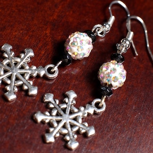 May include: A pair of silver snowflake earrings with a dangling design. Each earring features a detailed snowflake charm, a white, iridescent bead, and black beads. The earrings have silver-colored ear wires.