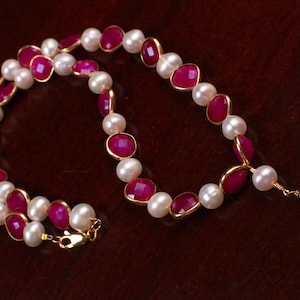 May include: A necklace made of white pearls and pink gemstones. The necklace has a drop pendant with a pink gemstone.