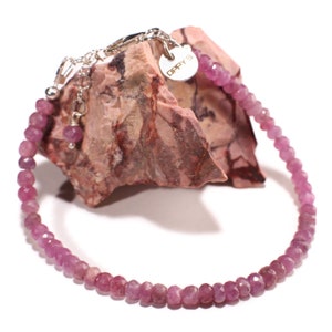 May include: A delicate pink gemstone bracelet with a silver clasp and a small charm that reads "OPPY'S". The bracelet is displayed on a rough, reddish-brown stone, showcasing the faceted beads.