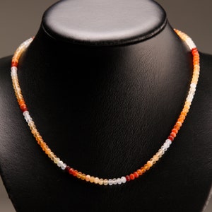 May include: A multi-colored opal gemstone necklace with white, orange, and red beads. The necklace is displayed on a black velvet background.