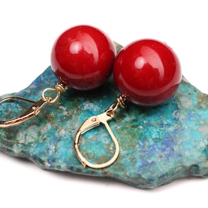 May include: A pair of red gemstone earrings with gold hoop style hooks. The earrings are displayed on a blue and green stone.