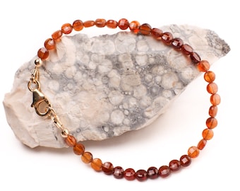 May include: A brown gemstone bracelet with a gold clasp. The bracelet is made of small, round beads and is lying on a light brown rock.
